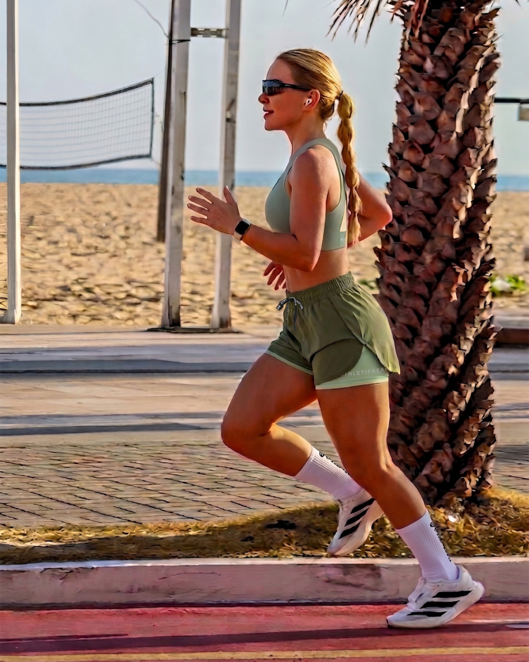 Woman running on a beach path with palm trees and a volleyball net in the background.