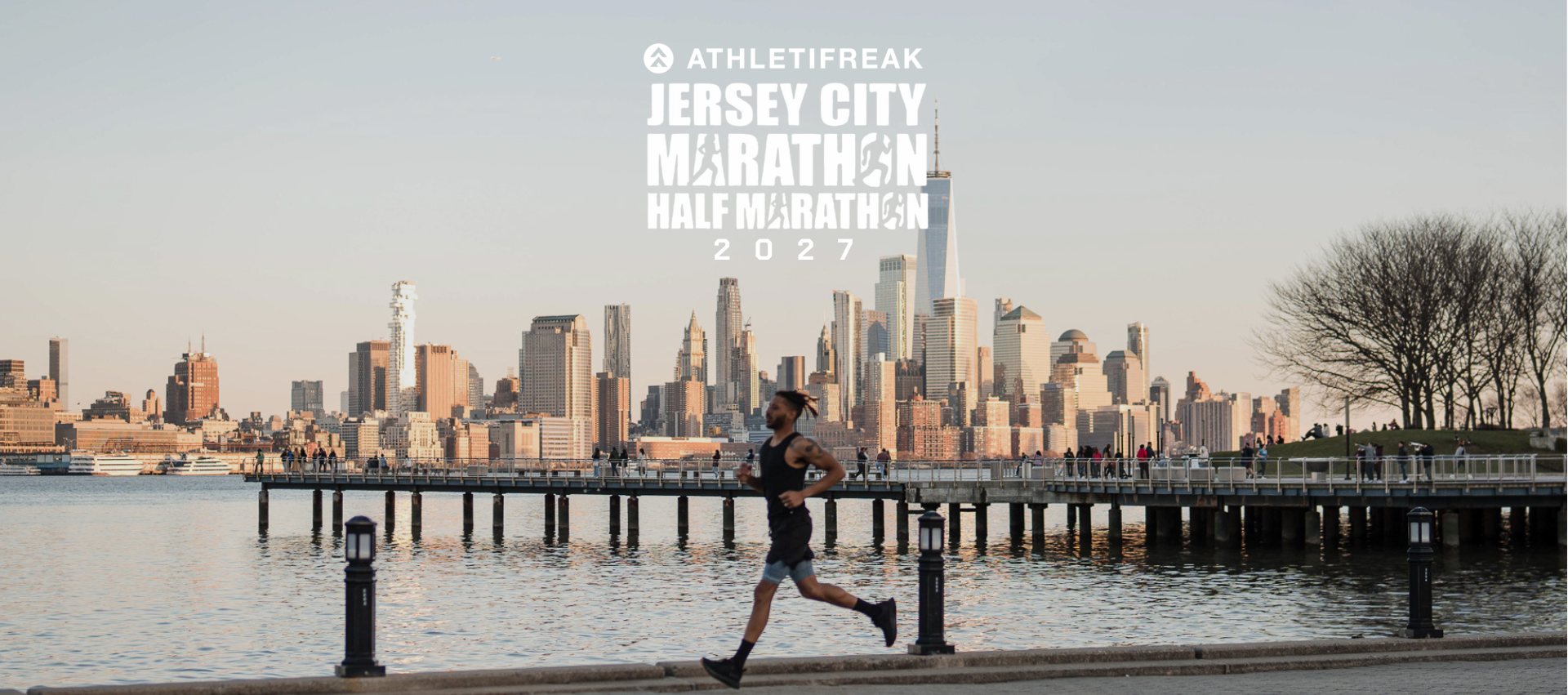 A runner strides along a waterfront promenade with the New York City skyline and a pier in the background. White overlaid text at the top reads 'ATHLETIFREAK JERSEY CITY MARATHON HALF MARATHON 2022'.