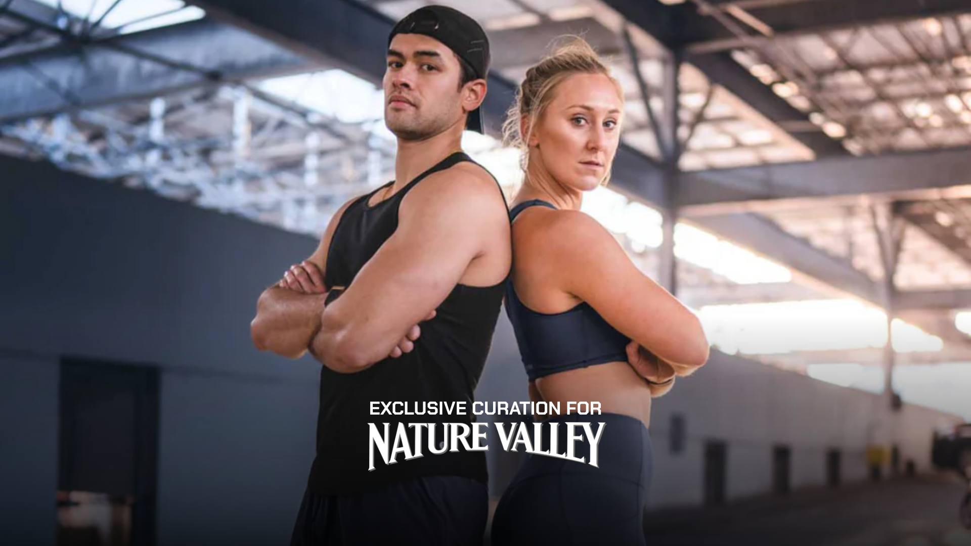 Two models, a man and a woman, standing back-to-back in a gym wearing athletic wear. The man wears a black tank top and black cap, while the woman wears a dark navy sports bra and matching leggings.