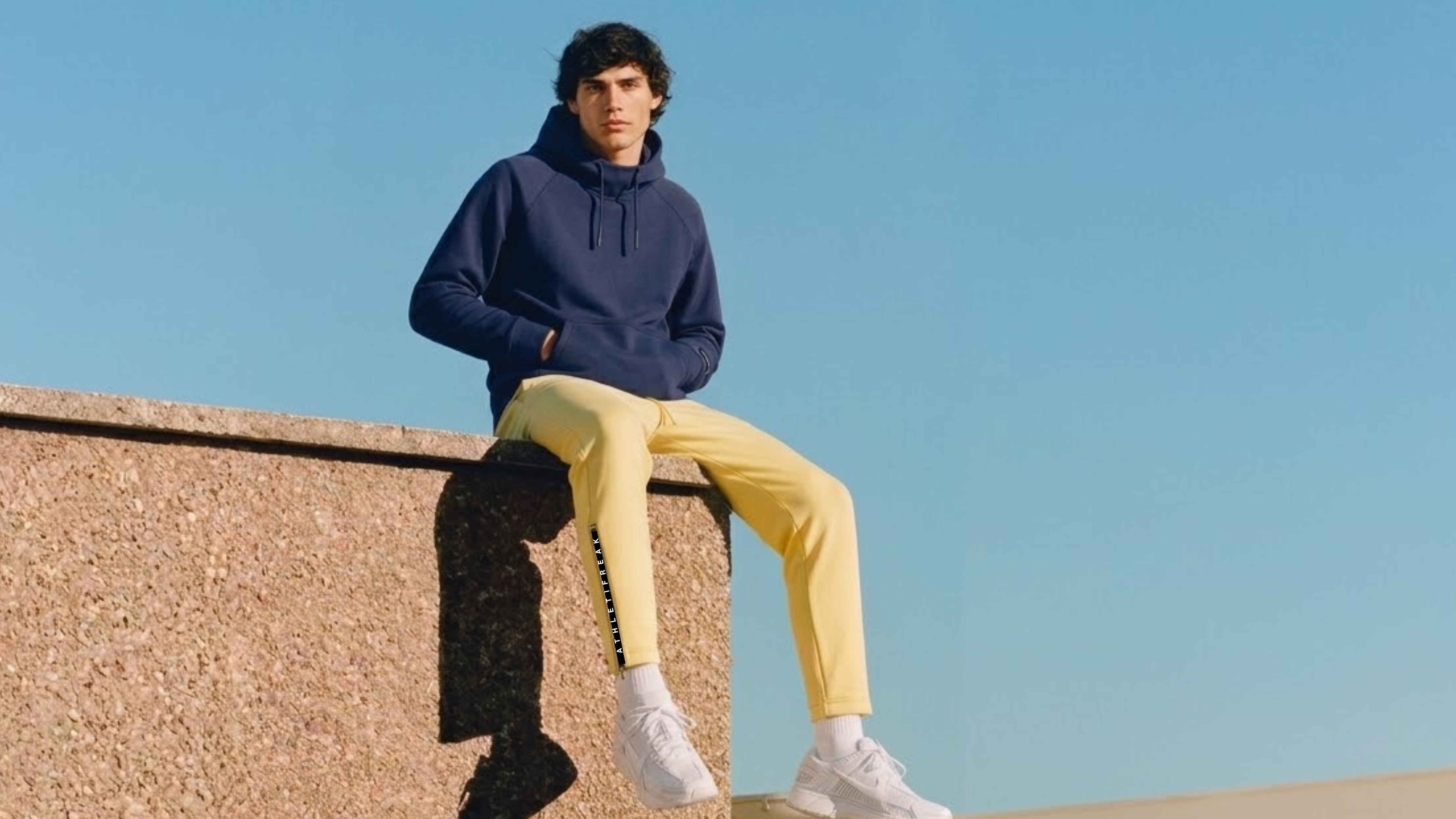 Person wearing a navy hoodie and yellow pants sitting on a ledge with a clear blue sky.